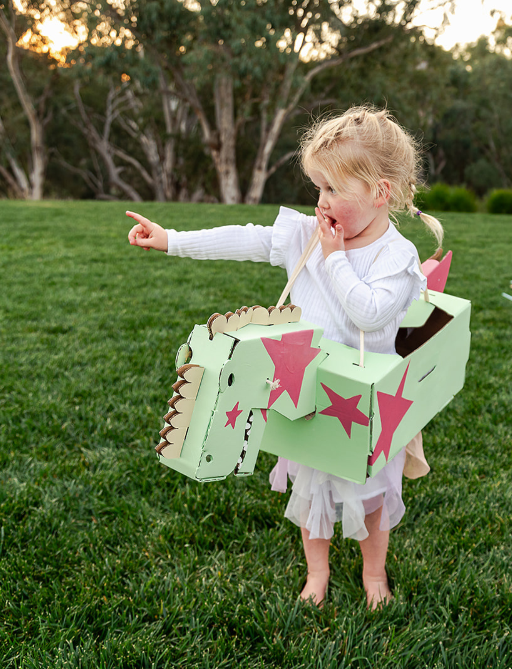 Two young children embark on prehistoric adventures with wearable cardboard animal costumes from Les Petite Artistes. The child on the left, in a pink sweater and blue jeans, holds a blue costume resembling a horse. The child on the right, wearing a beige sweater, joyfully brandishes their Nice Dinosaur Costume.