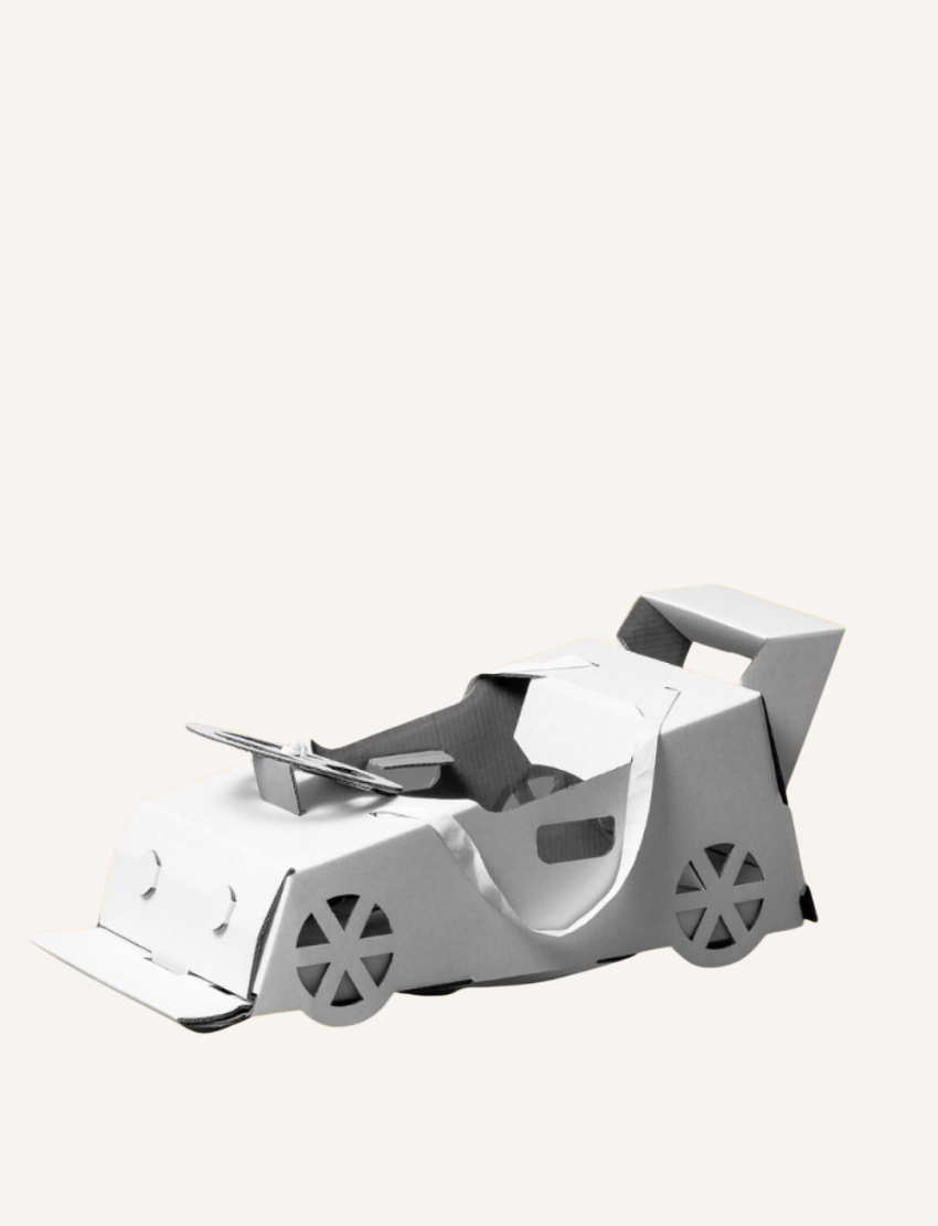 A small 3D model of a car made from white paper, featuring four wheels, a large rear wing, and cut-out windows. The paper's folds and creases define the car's shape and details. Named the "Race Car Costume" by Les Petite Artistes, it is set against a plain white background.