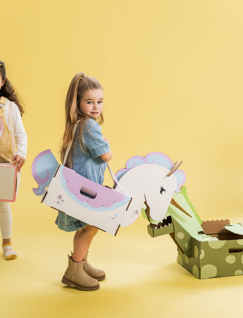 A person wearing a white shirt and a yellow apron is holding the Les Petite Artistes Unicorn Costume. The costume is adorned with pastel colors, including purple, blue, and a hint of pink, and features a handle for easy carrying.