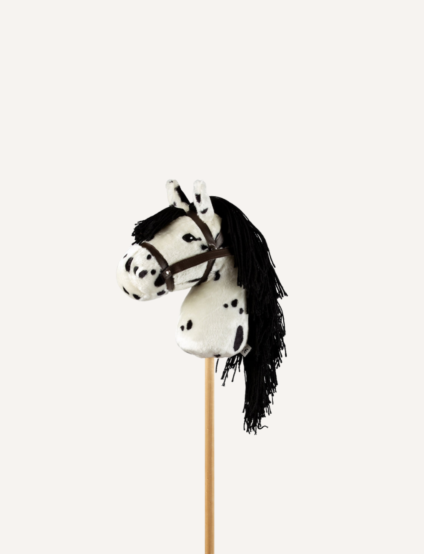 The Hobby Horse - Piebald by Astrup features a white and black spotted plush design with a black yarn mane, all mounted on a wooden stick, set against a plain white background.