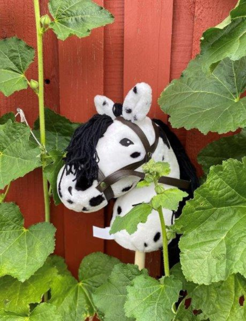 The Hobby Horse - Piebald by Astrup features a white and black spotted plush design with a black yarn mane, all mounted on a wooden stick, set against a plain white background.