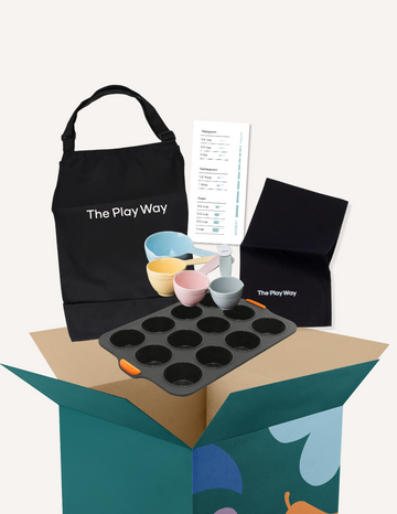 The "Getting Started with Baking Kit" by The Play Way is open. Inside this children's baking kit, you'll find a black apron and a black towel, both featuring "The Play Way" branding, colorful measuring cups, a recipe sheet for culinary adventures, and a black muffin tray.