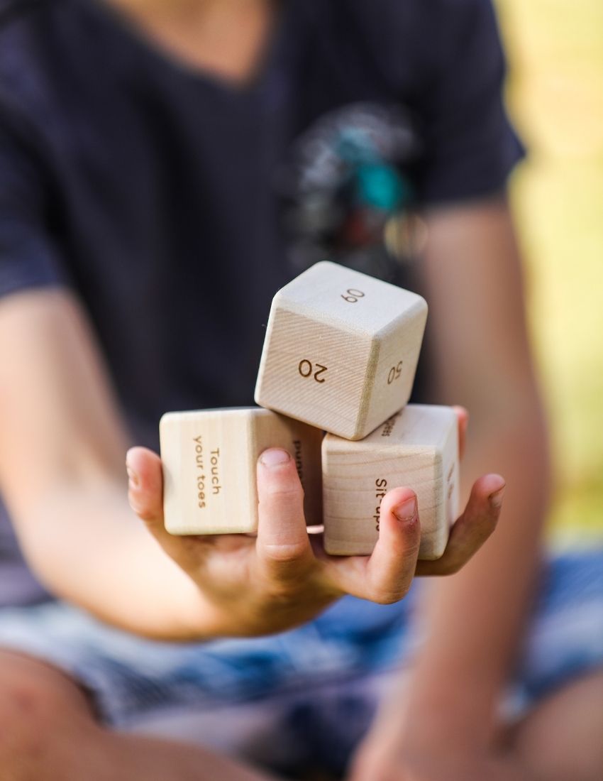 Introducing the Movement Dice Pack by The Play Way: a collection of wooden dice crafted for dynamic exercise routines, presented in a white cloth bag stamped with "The Play Way." Featuring instructions such as "Stand on one foot" alongside numerical values, these dice promote foundational strength through engaging play.