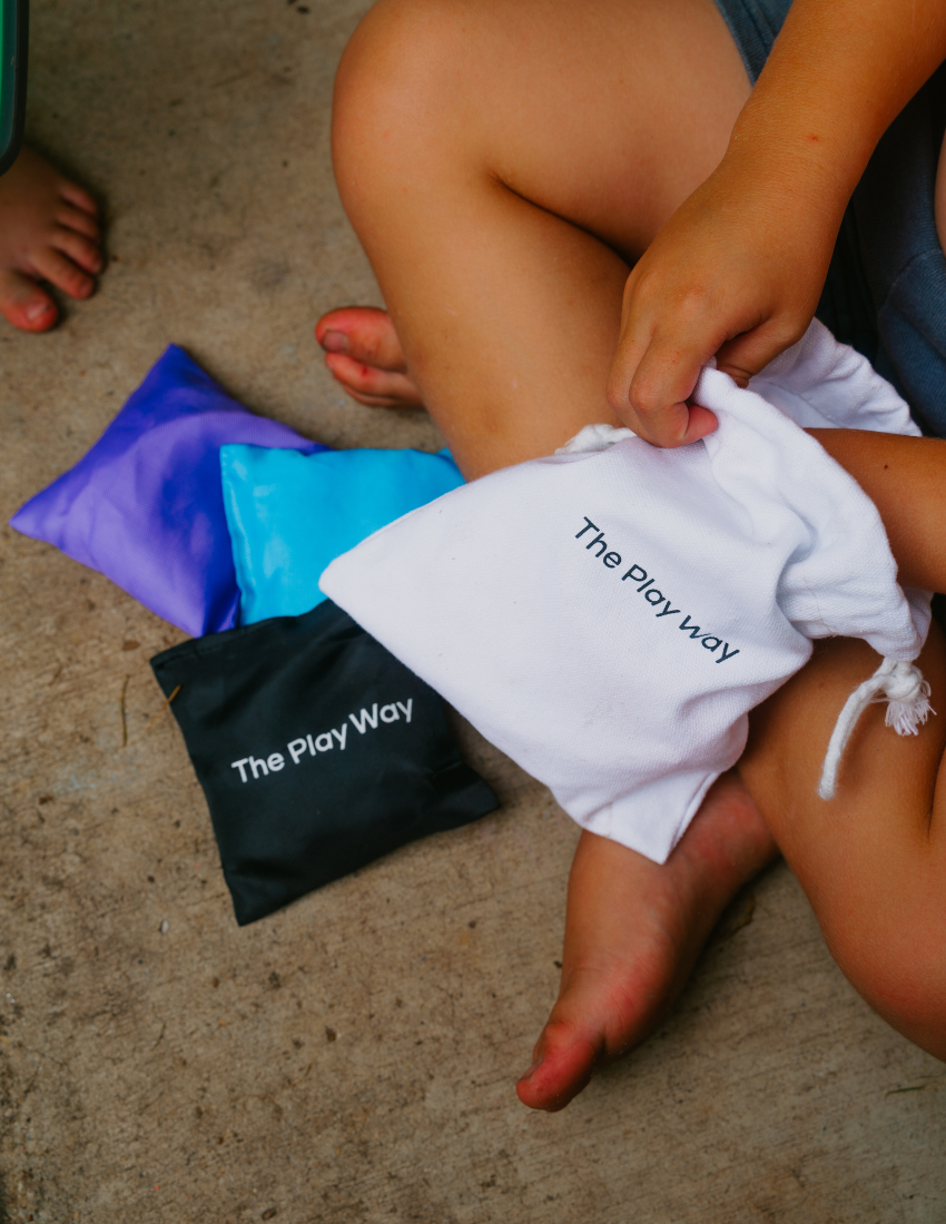 A beige drawstring bag labeled "Bean Bags and Ways to Play Booklet" from The Play Way brand sits beside a stack of colorful bean bags in orange, purple, green, blue, and yellow. A black bean bag with the same label completes the set. The small tag reads "The Play Way: An Indoor/Outdoor Fun Game," perfect for honing fine and gross motor skills.