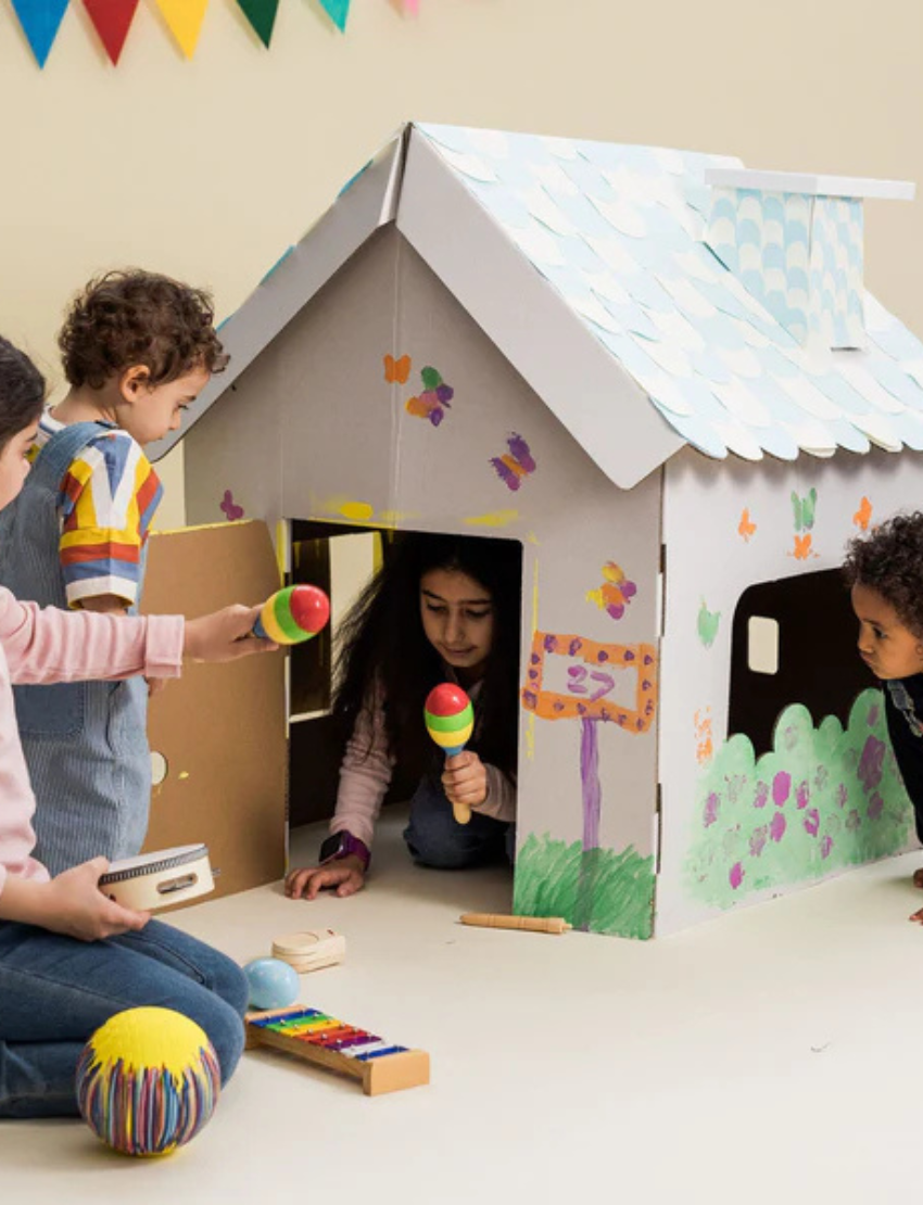 The Scalloped Cardboard House by Les Petite Artistes is a small white playhouse with a pitched roof and chimney. It features an open front with two window shutters and maintains a minimalist design, characterized by clean lines and a plain surface.