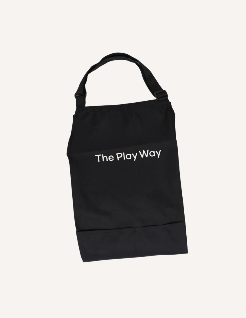 The "Getting Started with Baking Kit" by The Play Way is open. Inside this children's baking kit, you'll find a black apron and a black towel, both featuring "The Play Way" branding, colorful measuring cups, a recipe sheet for culinary adventures, and a black muffin tray.