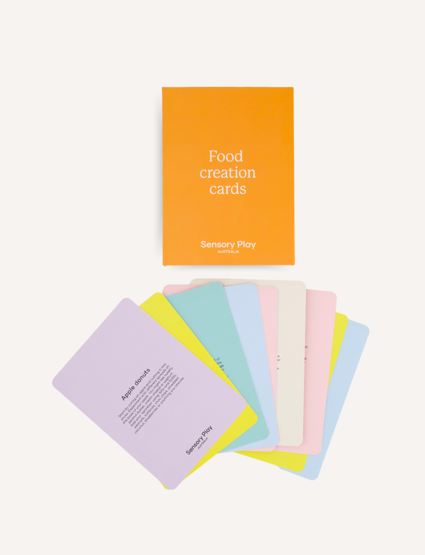 A teal and brown box from The Play Way's Food Exploration Kit, open to display its contents. Inside are a divided plate, utensils, a booklet titled "Food Creation Cards," two kitchen utensils, an orange pop-it toy for sensory preferences, a rolling pin set, and various cards labeled "Food Investigator." It's the ultimate Food Exploration Kit.