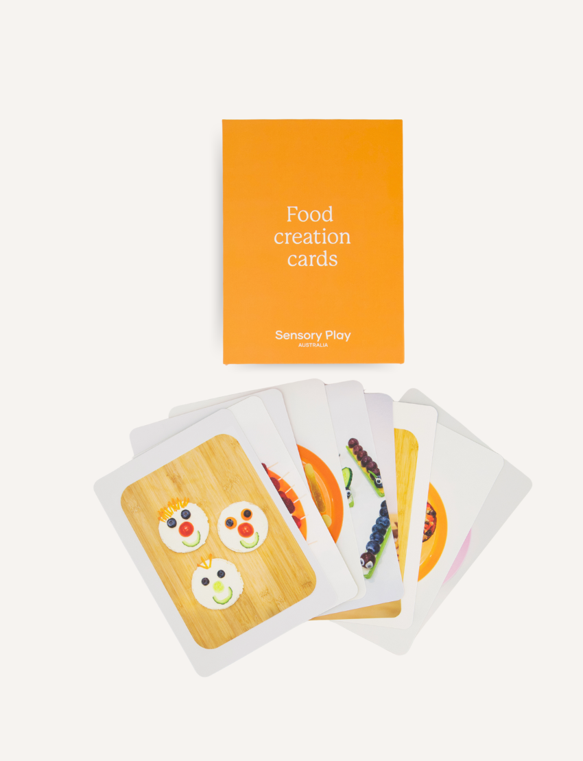 A teal and brown box from The Play Way's Food Exploration Kit, open to display its contents. Inside are a divided plate, utensils, a booklet titled "Food Creation Cards," two kitchen utensils, an orange pop-it toy for sensory preferences, a rolling pin set, and various cards labeled "Food Investigator." It's the ultimate Food Exploration Kit.