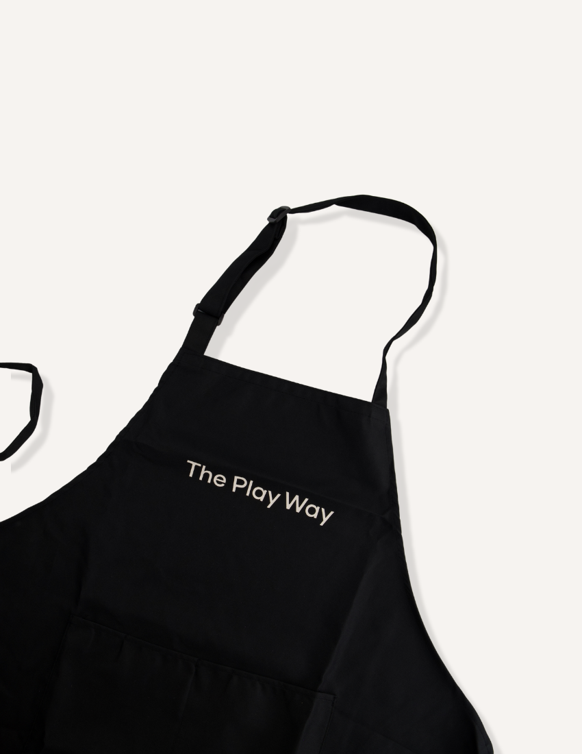 The "Getting Started with Baking Kit" by The Play Way is open. Inside this children's baking kit, you'll find a black apron and a black towel, both featuring "The Play Way" branding, colorful measuring cups, a recipe sheet for culinary adventures, and a black muffin tray.