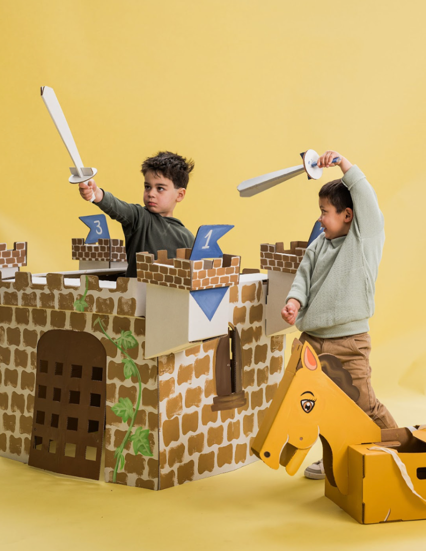 A cardboard pop-up castle with turrets unfolds from a green box, included in the Les Petite Artistes Noble Knight Kit. Designed by The Play Way x Les Petite Artistes, the castle comes complete with a drawbridge and includes a cardboard sword and shield. Accompanying it is a booklet titled "Noble Knight," adorned with a green sword design, offering an ideal backdrop for imaginative play and fostering cognitive development.