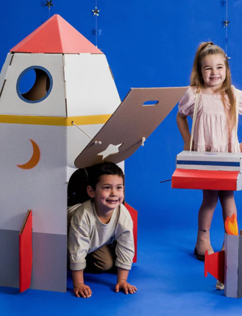 The Rocketship Playhouse by Les Petite Artistes features a large front door and cut-out shapes of a star and crescent moon on its sides. This primarily monochrome cardboard structure is designed as an imaginative play space for children.