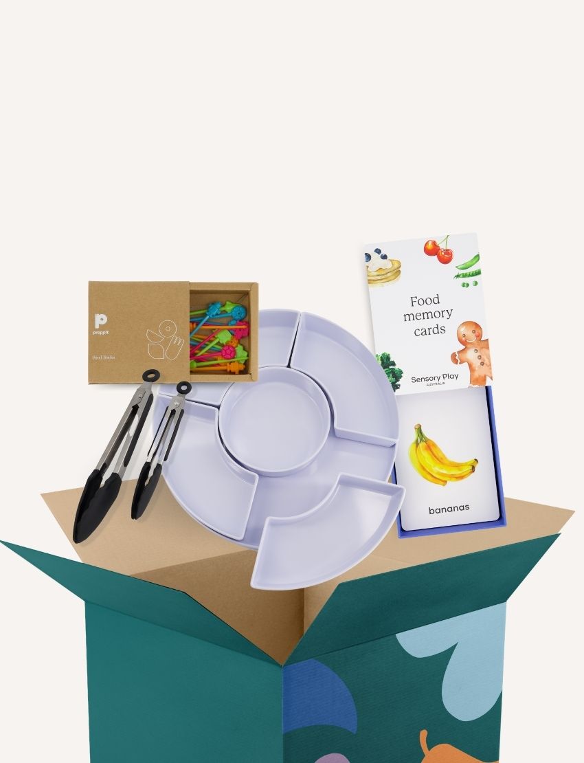 A green box, part of The Play Way's Food Sharing Kit, is filled with various items to enhance mealtime experiences. Inside, you'll find a set of tongs, a silicone tray with five compartments, colorful rubber bands, and a pack of food memory cards featuring an image of bananas for sensory needs.