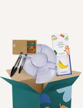 The Play Way's Family Meal and Food Sharing Kit features a cardboard box with a colorful design that opens to reveal various items, including a compartmentalized plastic tray, two sculpting tools, a set of food memory cards for sensory play, and a small box containing multicolored rubber bands.