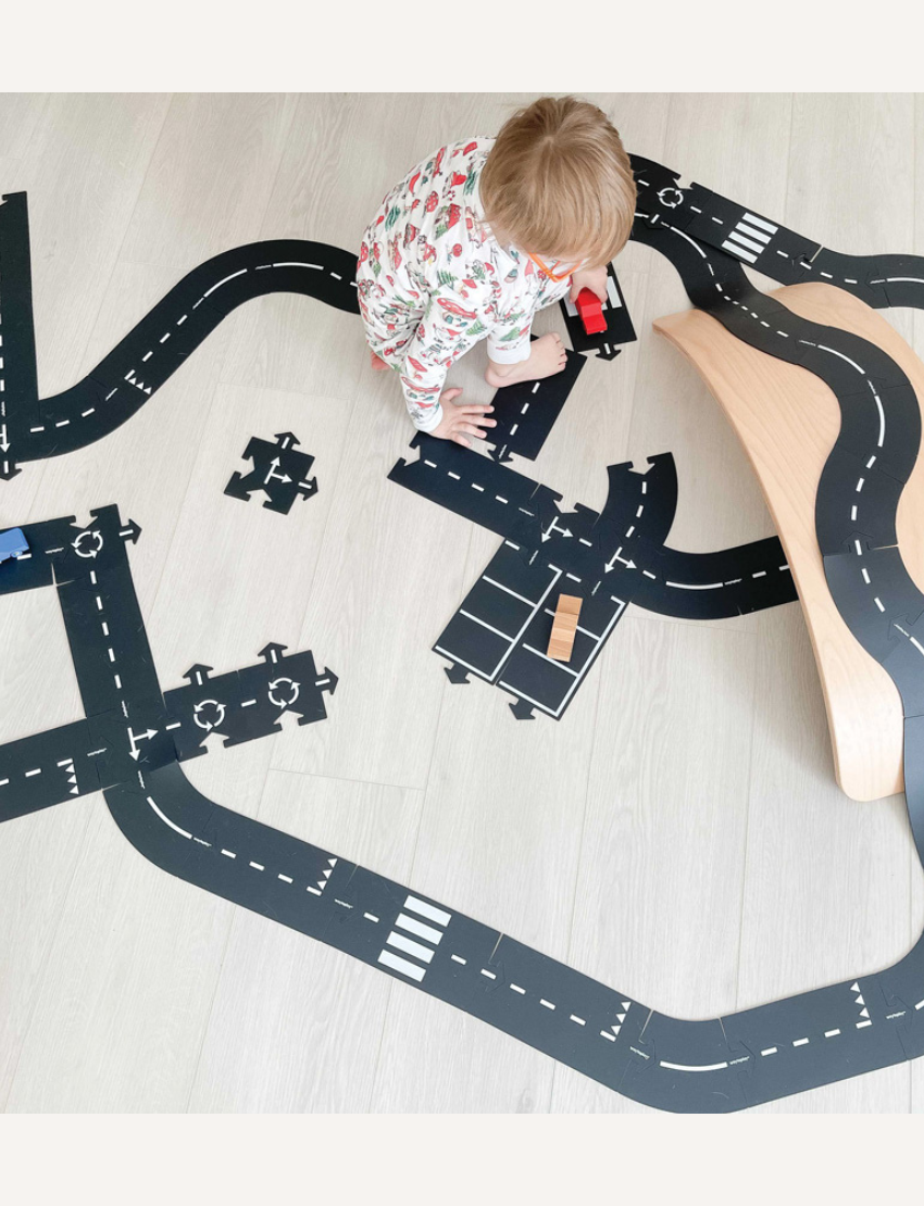 The image displays the "King Of The Road Car Edition" toy set by Waytoplay, which includes flexible road pieces and two wooden cars in red and blue. In the background, there is a box depicting a child playing with and assembling the road.