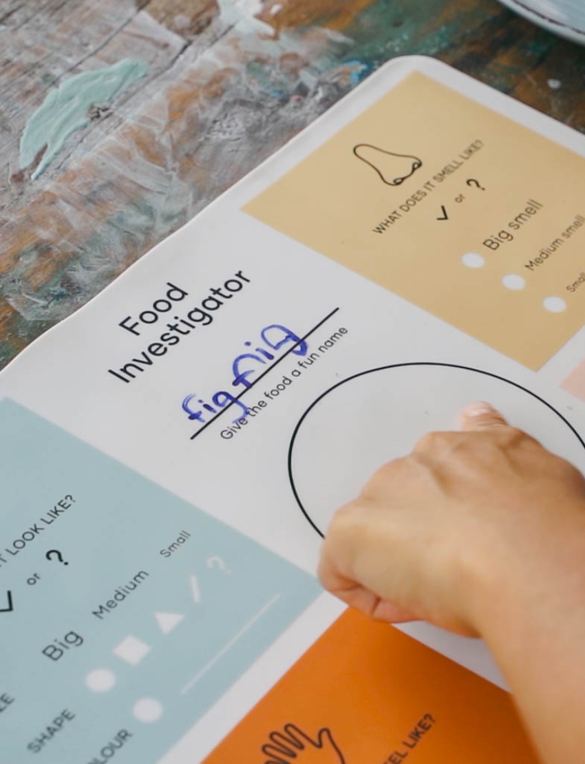 A teal and brown box from The Play Way's Food Exploration Kit, open to display its contents. Inside are a divided plate, utensils, a booklet titled "Food Creation Cards," two kitchen utensils, an orange pop-it toy for sensory preferences, a rolling pin set, and various cards labeled "Food Investigator." It's the ultimate Food Exploration Kit.