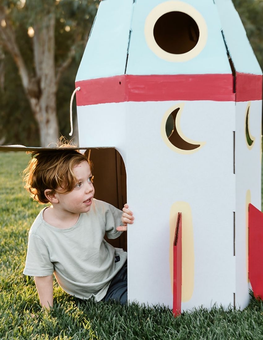 A green box opens to reveal the "Les Petite Artistes Space Kit" by The Play Way, featuring a gray cardboard rocket ship, various space-themed cardboard cutouts, and a booklet titled "Space" adorned with a rocket illustration on its cover. This imaginative play kit provides playful and educational elements for endless space-themed adventures.