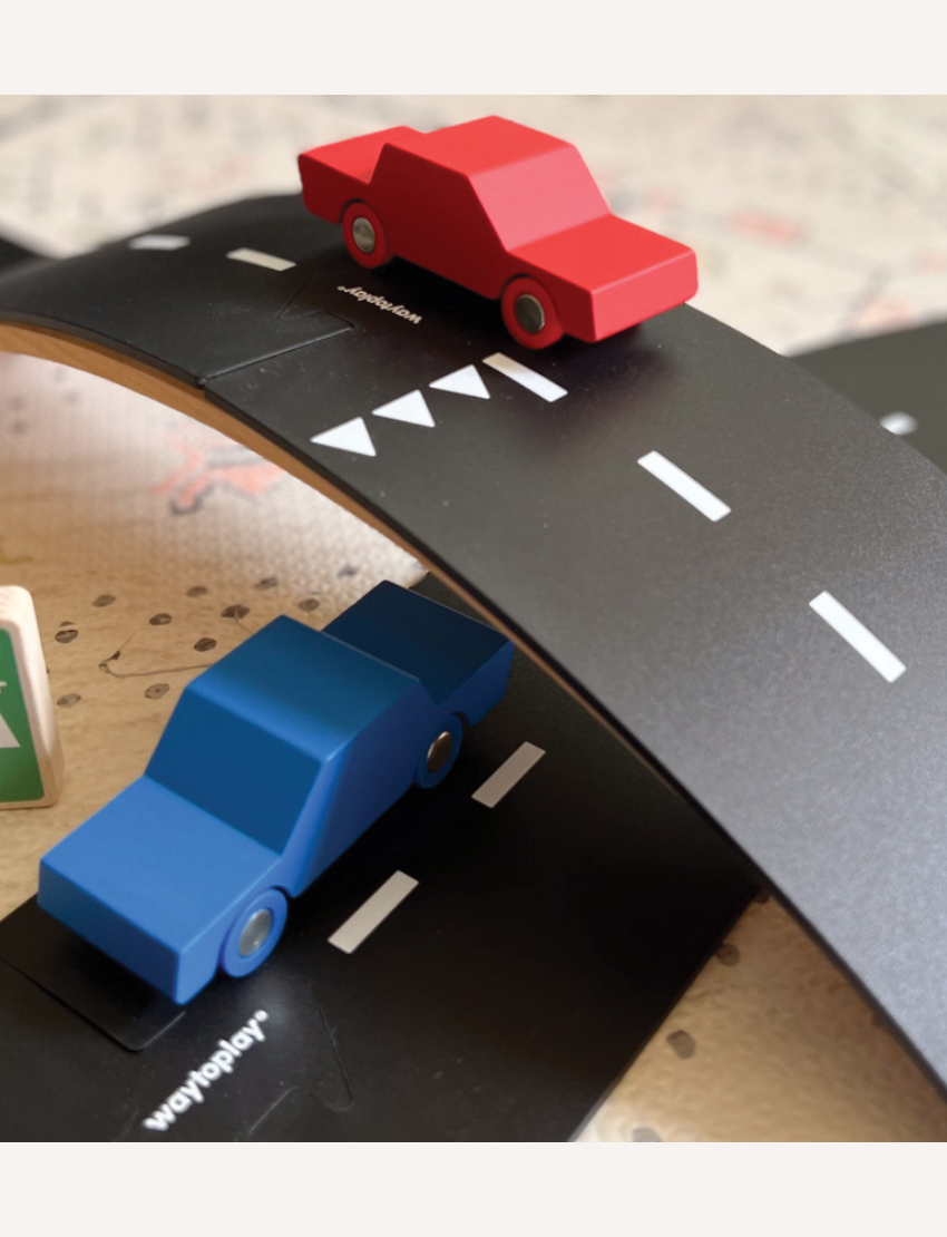 The image displays the "King Of The Road Car Edition" toy set by Waytoplay, which includes flexible road pieces and two wooden cars in red and blue. In the background, there is a box depicting a child playing with and assembling the road.