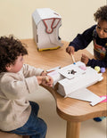 A colorful cardboard box from The Play Way contains a Space Kit, perfect for engaging in space-themed activities. Inside, assembled cardboard pieces form a rocket, a small spaceship, and an astronaut helmet. A booklet titled 