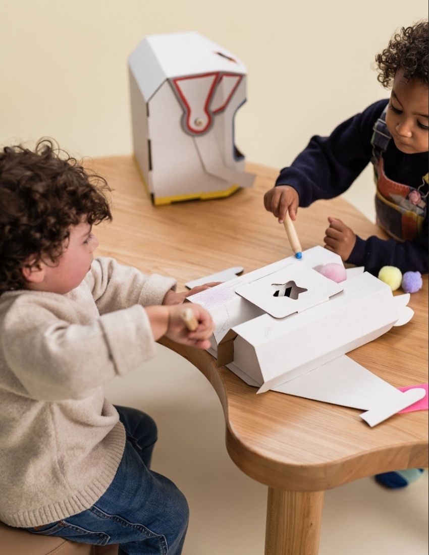 A colorful cardboard box from The Play Way contains a Space Kit, perfect for engaging in space-themed activities. Inside, assembled cardboard pieces form a rocket, a small spaceship, and an astronaut helmet. A booklet titled "Space," adorned with a rocket illustration, is included at the front of the kit to facilitate imaginative play.