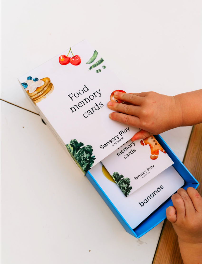 The Play Way's Family Meal and Food Sharing Kit features a cardboard box with a colorful design that opens to reveal various items, including a compartmentalized plastic tray, two sculpting tools, a set of food memory cards for sensory play, and a small box containing multicolored rubber bands.