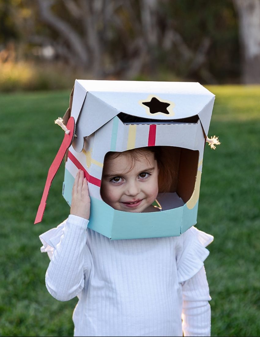 A colorful cardboard box from The Play Way contains a Space Kit, perfect for engaging in space-themed activities. Inside, assembled cardboard pieces form a rocket, a small spaceship, and an astronaut helmet. A booklet titled "Space," adorned with a rocket illustration, is included at the front of the kit to facilitate imaginative play.