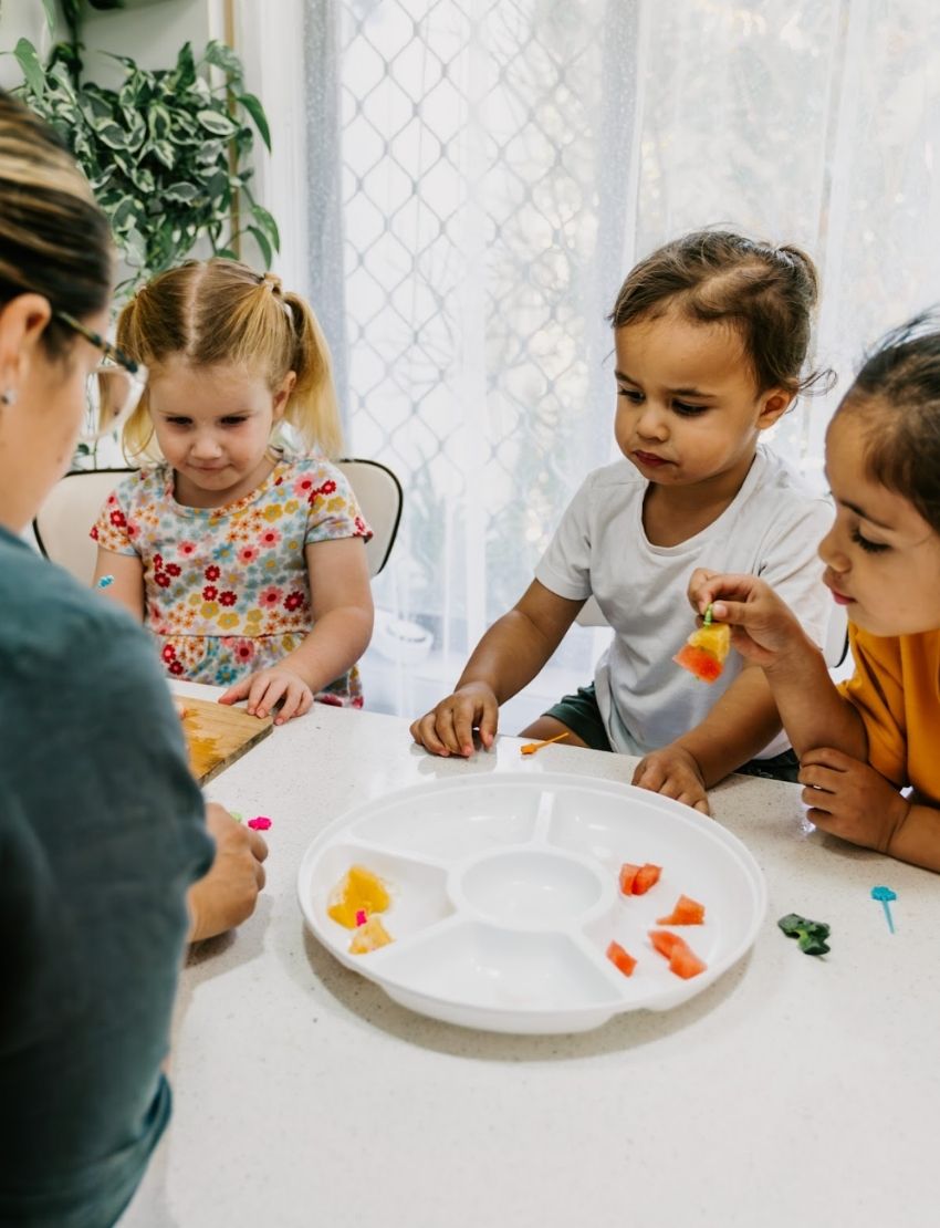 A green box, part of The Play Way's Food Sharing Kit, is filled with various items to enhance mealtime experiences. Inside, you'll find a set of tongs, a silicone tray with five compartments, colorful rubber bands, and a pack of food memory cards featuring an image of bananas for sensory needs.