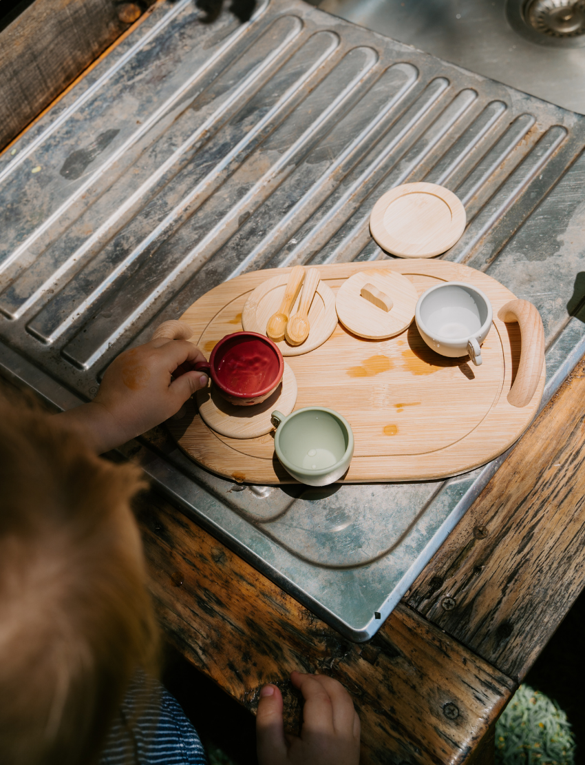 A child indulges in imaginative play with Sensory Play Australia's wooden Teatime Set, featuring a tray, three cups, a teapot, and spoons. Nearby is a metal sink with a draining area on the countertop to enhance cognitive skills through interactive fun.