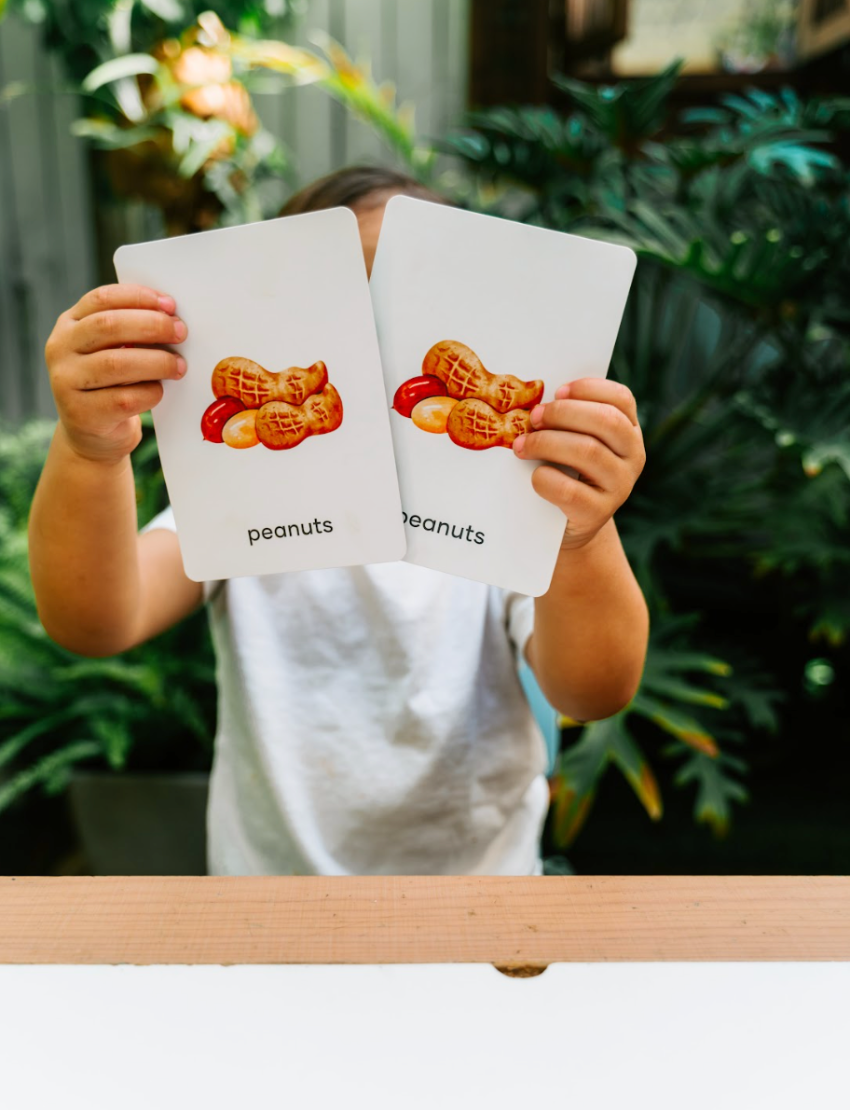 The Play Way's Family Meal and Food Sharing Kit features a cardboard box with a colorful design that opens to reveal various items, including a compartmentalized plastic tray, two sculpting tools, a set of food memory cards for sensory play, and a small box containing multicolored rubber bands.