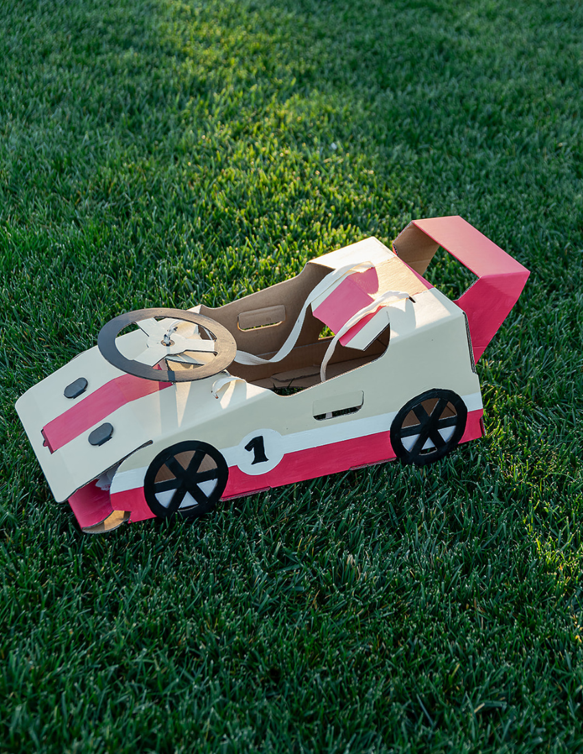 A cardboard box is open, showcasing assembled white and gray paper models of futuristic vehicles like cars and motorcycles. A small orange card labeled "Les Petitie Artistes Transport Kit" from The Play Way accompanies the models, encouraging imaginative play and turning each vehicle into a gateway for transport-themed adventures.