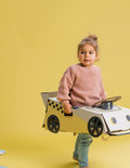 A large teal and orange cardboard box from The Play Way's Transport Kit is open, revealing paper models of vintage vehicles inside. The kit includes models of a carriage, an automobile, and a small cart. A card featuring the word 