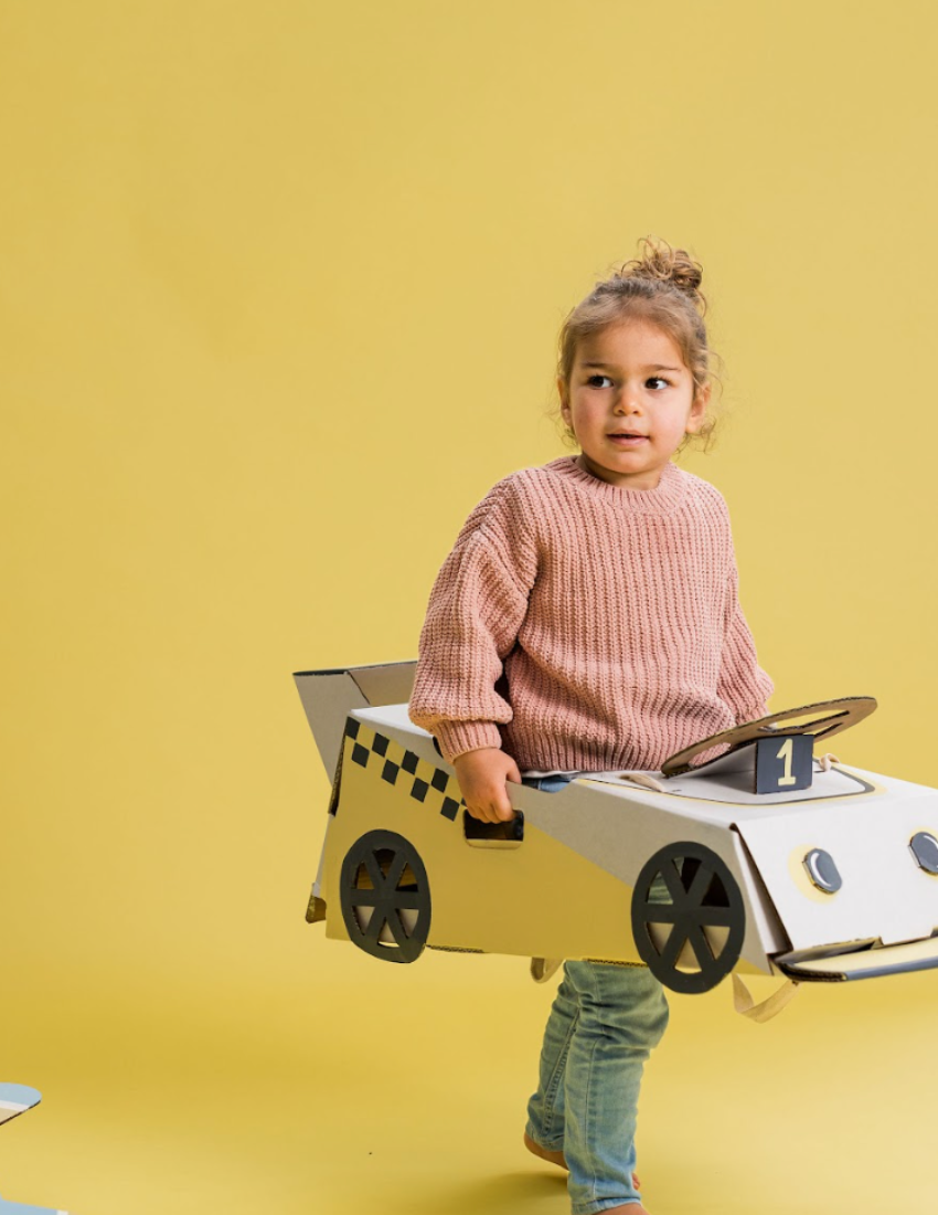 A large teal and orange cardboard box from The Play Way's Transport Kit is open, revealing paper models of vintage vehicles inside. The kit includes models of a carriage, an automobile, and a small cart. A card featuring the word "Transport" along with an illustration of a boat enhances the imaginative play experience.