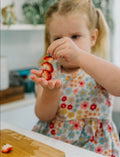 The Play Way's Family Meal and Food Sharing Kit features a cardboard box with a colorful design that opens to reveal various items, including a compartmentalized plastic tray, two sculpting tools, a set of food memory cards for sensory play, and a small box containing multicolored rubber bands.