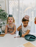 The Play Way's Family Meal and Food Sharing Kit features a cardboard box with a colorful design that opens to reveal various items, including a compartmentalized plastic tray, two sculpting tools, a set of food memory cards for sensory play, and a small box containing multicolored rubber bands.