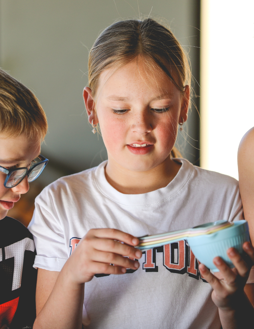 The "Getting Started with Baking Kit" by The Play Way is open. Inside this children's baking kit, you'll find a black apron and a black towel, both featuring "The Play Way" branding, colorful measuring cups, a recipe sheet for culinary adventures, and a black muffin tray.