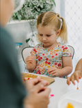 The Play Way's Family Meal and Food Sharing Kit features a cardboard box with a colorful design that opens to reveal various items, including a compartmentalized plastic tray, two sculpting tools, a set of food memory cards for sensory play, and a small box containing multicolored rubber bands.