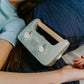 A parent with two kids is on a bed. They and one child are exploring The Play Way's Sleep Program box, while the other child holds a book. The bedroom features a decorative headboard and a patterned lamp.