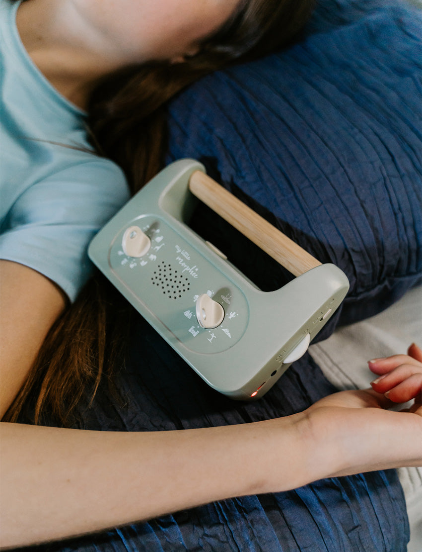 A parent with two kids is on a bed. They and one child are exploring The Play Way's Sleep Program box, while the other child holds a book. The bedroom features a decorative headboard and a patterned lamp.