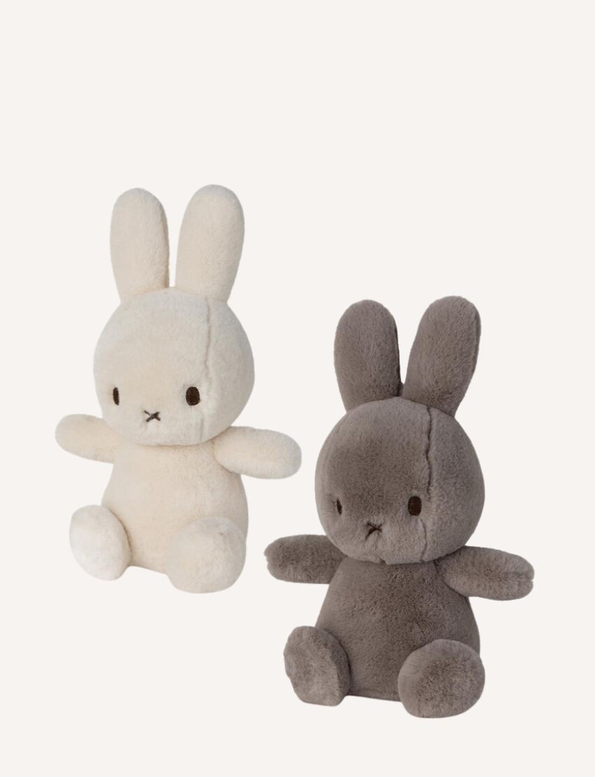 Two Miffy "Cozy Miffy" plush bunnies are showcased against a plain background. The left one is light cream, while the right is dark gray; both are made from recycled polyester and feature simple facial details with small black eyes and a cross-shaped nose and mouth.