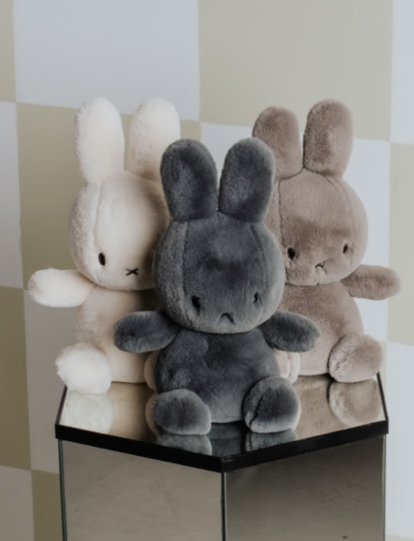 Two Miffy "Cozy Miffy" plush bunnies are showcased against a plain background. The left one is light cream, while the right is dark gray; both are made from recycled polyester and feature simple facial details with small black eyes and a cross-shaped nose and mouth.