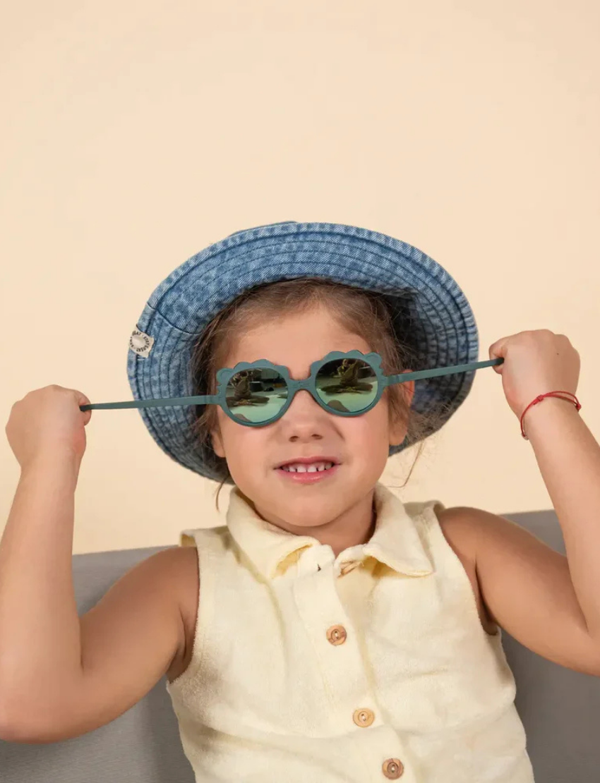 Two pairs of Ki ET LA's LION sunglasses for 2-4-year-olds are displayed. One, made in France, has dark frames with blue-tinted lenses; the other features light yellow frames with gold-tinted lenses. Both have round, unbreakable lenses and decorative frame accents.