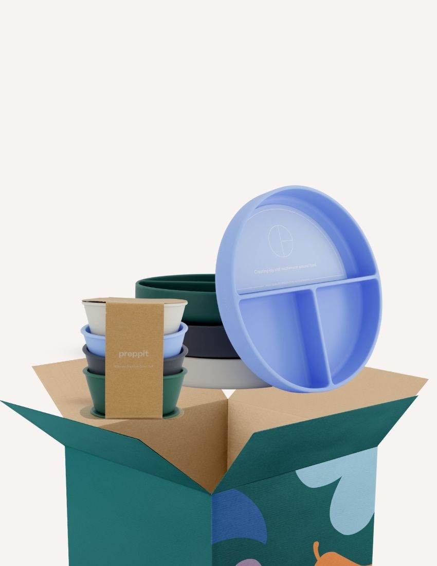 An open green and teal cardboard box from The Play Way's Tools For Eating - Bowls & Plates Kit contains a set of stackable silicone suction bowls, a set of stackable cups, and an upright blue divided baby plate. The items are neatly arranged, showcasing their self-feeding design.