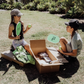 Two children sit on grass by an open cardboard box from The Play Way's "Building Motor Skills through Play Program," featuring various items. Holding green paddles with text, they wear hats and casual clothes. Bushes and trees in the background suggest a park setting.