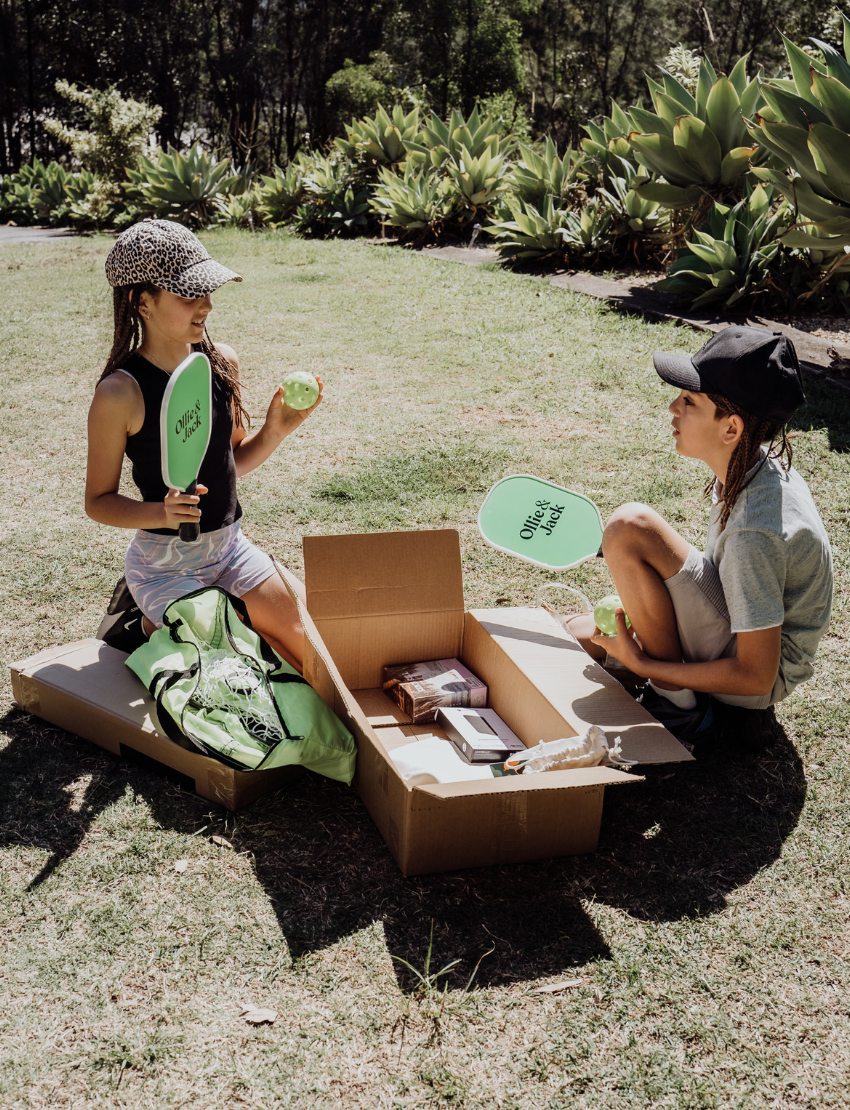 Two children sit on grass by an open cardboard box from The Play Way's "Building Motor Skills through Play Program," featuring various items. Holding green paddles with text, they wear hats and casual clothes. Bushes and trees in the background suggest a park setting.