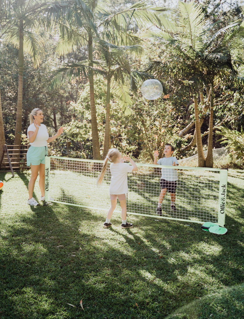 Two children sit on grass by an open cardboard box from The Play Way's "Building Motor Skills through Play Program," featuring various items. Holding green paddles with text, they wear hats and casual clothes. Bushes and trees in the background suggest a park setting.