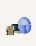 An open green and teal cardboard box from The Play Way's Tools For Eating - Bowls & Plates Kit contains a set of stackable silicone suction bowls, a set of stackable cups, and an upright blue divided baby plate. The items are neatly arranged, showcasing their self-feeding design.