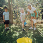 Two children sit on grass by an open cardboard box from The Play Way's "Building Motor Skills through Play Program," featuring various items. Holding green paddles with text, they wear hats and casual clothes. Bushes and trees in the background suggest a park setting.