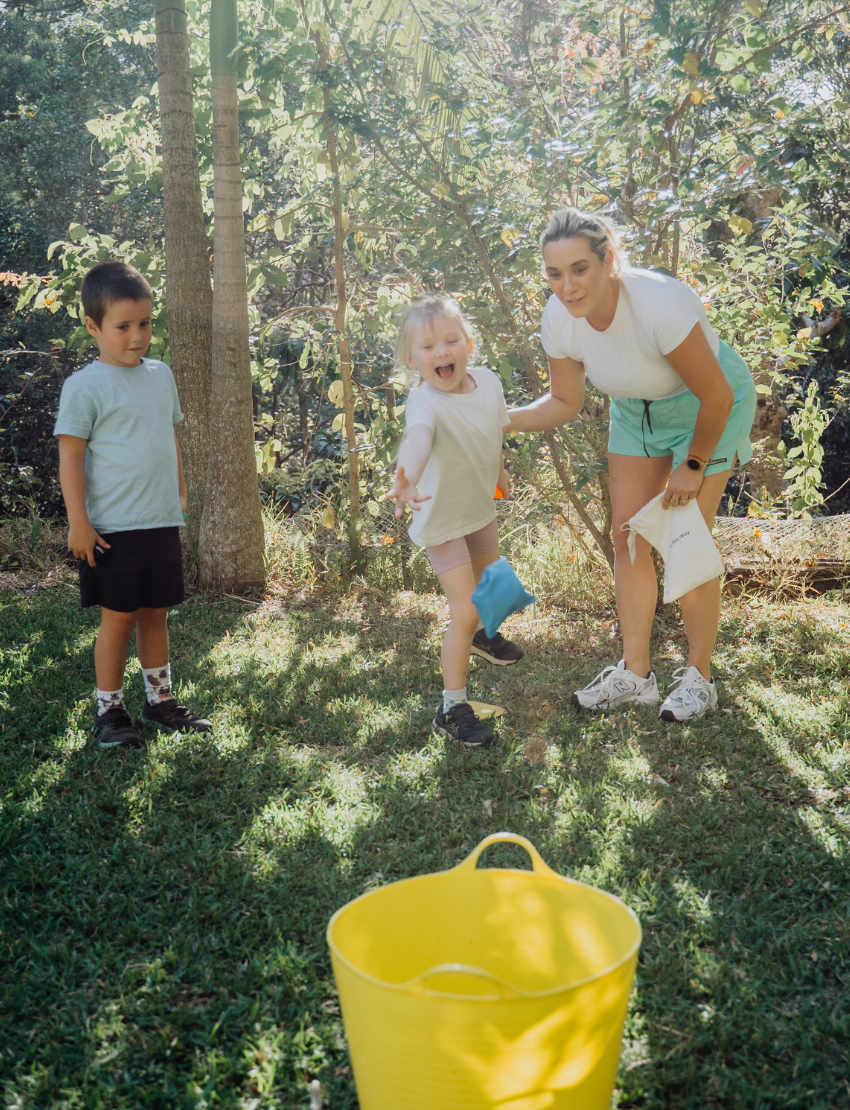 Two children sit on grass by an open cardboard box from The Play Way's "Building Motor Skills through Play Program," featuring various items. Holding green paddles with text, they wear hats and casual clothes. Bushes and trees in the background suggest a park setting.