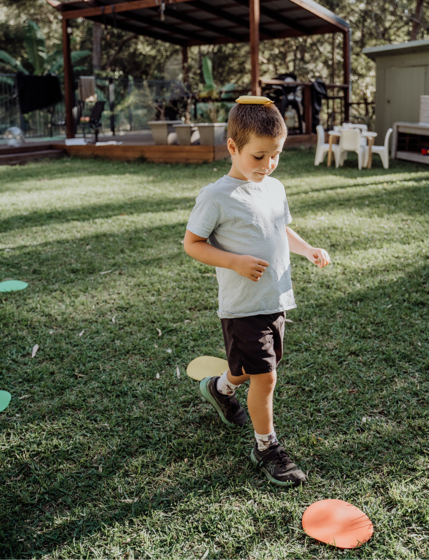Two children sit on grass by an open cardboard box from The Play Way's "Building Motor Skills through Play Program," featuring various items. Holding green paddles with text, they wear hats and casual clothes. Bushes and trees in the background suggest a park setting.
