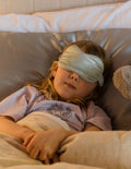 A child with blonde hair is lying in bed, wearing a light blue sleep mask over her eyes. She is dressed in a light purple shirt and tucked in under a beige blanket. Next to her, a stuffed animal is partially visible. She might be dreaming about tomorrow's kids' lunches prepared with The Play Way Lunch Buddy Planning Tools Set.