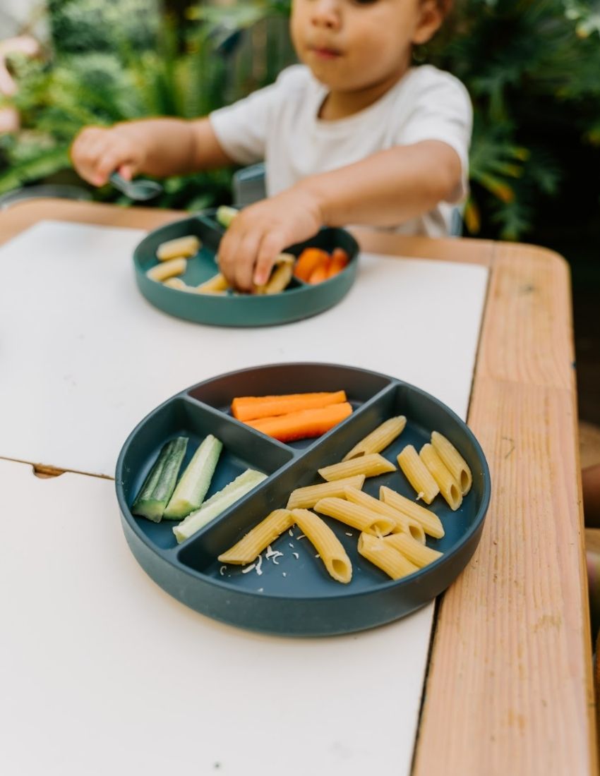 An open green and teal cardboard box from The Play Way's Tools For Eating - Bowls & Plates Kit contains a set of stackable silicone suction bowls, a set of stackable cups, and an upright blue divided baby plate. The items are neatly arranged, showcasing their self-feeding design.